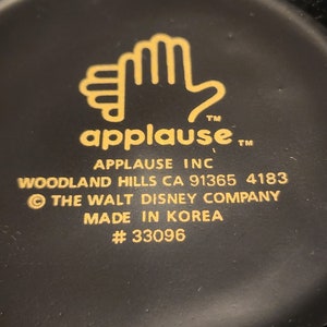 May include: Black and gold mug with a hand logo and the text "applause" and "APPLAUSE INC". The mug also includes the text "WOODLAND HILLS CA 91365 4183", "&copy; THE WALT DISNEY COMPANY", "MADE IN KOREA", and "#33096".