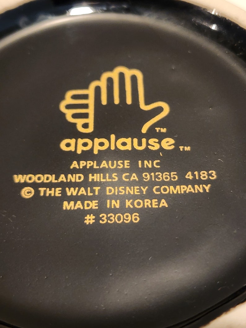 May include: Black and gold mug with a hand logo and the text "applause" and "APPLAUSE INC". The mug also includes the text "WOODLAND HILLS CA 91365 4183", "&copy; THE WALT DISNEY COMPANY", "MADE IN KOREA", and "#33096".