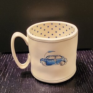 K&ouml;nnte beinhalten: Wei&szlig;e Keramiktasse mit einer blauen Autoillustration und einem blauen Tupfenmuster im Inneren. Die Tasse hat einen gebogenen Henkel und einen blauen Rand. Das Auto ist hellblau mit einer Flagge.
