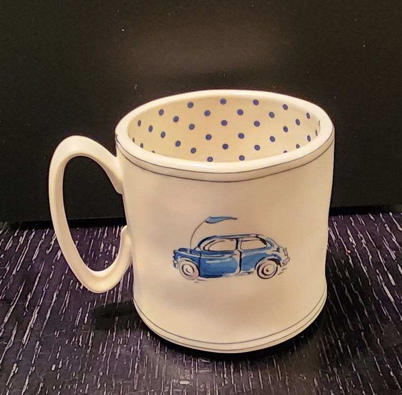 K&ouml;nnte beinhalten: Wei&szlig;e Keramiktasse mit einer blauen Autoillustration und einem blauen Tupfenmuster im Inneren. Die Tasse hat einen gebogenen Henkel und einen blauen Rand. Das Auto ist hellblau mit einer Flagge.