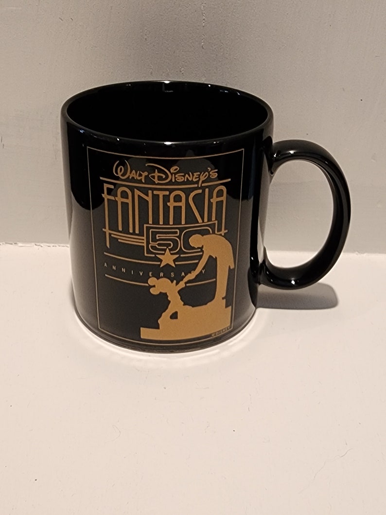 May include: Black ceramic coffee mug with a gold design featuring Mickey Mouse and the text "Walt Disney's Fantasia 50th Anniversary".