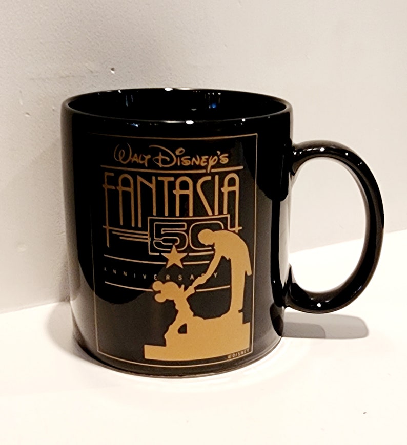 May include: Black ceramic coffee mug with a gold design featuring Mickey Mouse and Walt Disney's Fantasia 50th Anniversary logo.