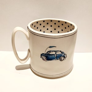 K&ouml;nnte beinhalten: Eine wei&szlig;e Keramik-Tasse mit schwarzem Henkel und dem Text "But First, Coffee" in fetter, schwarzer Schrift. Die Tasse steht vor einem verschwommenen Hintergrund, der eine gem&uuml;tliche Umgebung suggeriert.