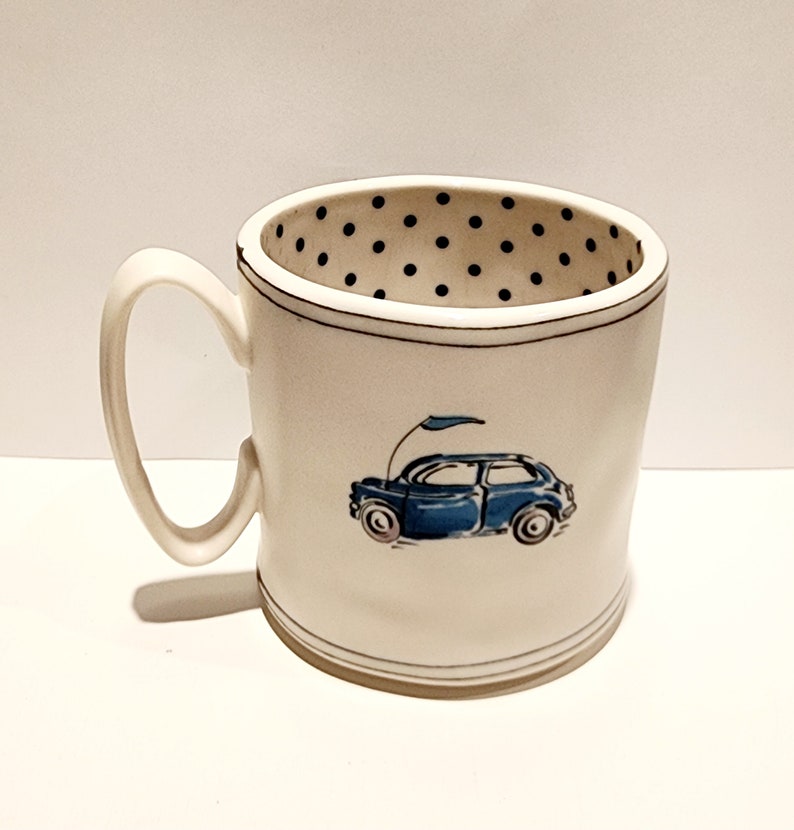 K&ouml;nnte beinhalten: Eine wei&szlig;e Keramik-Tasse mit schwarzem Henkel und dem Text "But First, Coffee" in fetter, schwarzer Schrift. Die Tasse steht vor einem verschwommenen Hintergrund, der eine gem&uuml;tliche Umgebung suggeriert.
