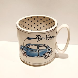 K&ouml;nnte beinhalten: Wei&szlig;e Keramiktasse mit einer blauen Autoillustration und dem Schriftzug "Bon Voyage". Das Innere der Tasse hat ein schwarzes Tupfenmuster. Die Tasse hat einen gebogenen Griff.
