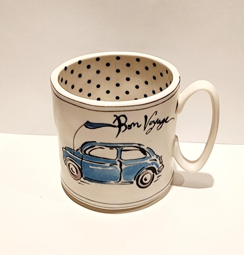 K&ouml;nnte beinhalten: Wei&szlig;e Keramiktasse mit einer blauen Autoillustration und dem Schriftzug "Bon Voyage". Das Innere der Tasse hat ein schwarzes Tupfenmuster. Die Tasse hat einen gebogenen Griff.