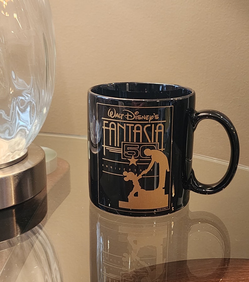 May include: Black ceramic coffee mug with a gold design featuring Mickey Mouse and the text "Walt Disney's Fantasia 50th Anniversary".