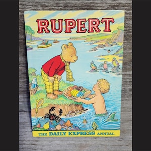 Puede incluir: Una portada de libro vintage de Rupert el Oso con una ilustraci&oacute;n colorida de Rupert el Oso con una camisa roja y pantalones amarillos, sosteniendo una cesta de fruta. La portada tambi&eacute;n presenta una sirena con cola, un cangrejo y una variedad de p&aacute;jaros. El texto en la portada dice "Rupert" y "The Daily Express Annual".