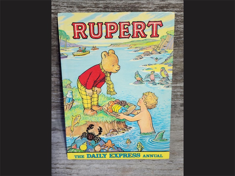 Puede incluir: Una portada de libro vintage de Rupert el Oso con una ilustraci&oacute;n colorida de Rupert el Oso con una camisa roja y pantalones amarillos, sosteniendo una cesta de fruta. La portada tambi&eacute;n presenta una sirena con cola, un cangrejo y una variedad de p&aacute;jaros. El texto en la portada dice "Rupert" y "The Daily Express Annual".