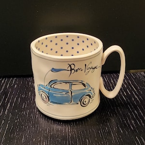 K&ouml;nnte beinhalten: Wei&szlig;e Keramiktasse mit einer blauen Autoillustration und dem Schriftzug "Bon Voyage". Der Innenraum weist ein blaues Tupfenmuster auf. Die Tasse hat einen gebogenen Henkel und steht vor einem dunklen Hintergrund.