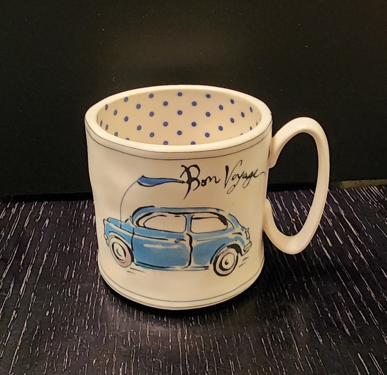 K&ouml;nnte beinhalten: Wei&szlig;e Keramiktasse mit einer blauen Autoillustration und dem Schriftzug "Bon Voyage". Der Innenraum weist ein blaues Tupfenmuster auf. Die Tasse hat einen gebogenen Henkel und steht vor einem dunklen Hintergrund.
