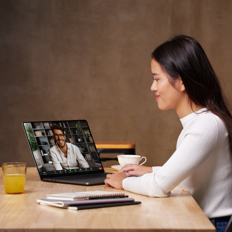May include: A woman in a white sweater sits at a wooden table, looking at a laptop screen. The screen displays a video call with a smiling man. A glass of orange juice, a cup of coffee, and notebooks are on the table.
