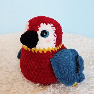 Può includere: Un pappagallo di peluche fatto a mano dai colori vivaci. L'uccello presenta una testa e un corpo rossi, ali blu, un becco nero e dettagli bianchi intorno agli occhi. Il pappagallo è seduto su una superficie bianca e soffice.