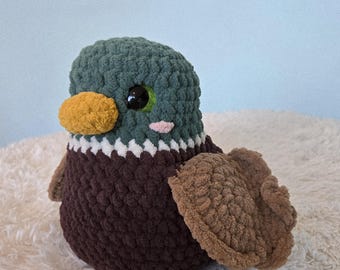 Puede incluir: Un peluche de pato hecho a mano con ganchillo, con cabeza verde azulado, cuerpo marrón y alas color canela. El pato tiene un pico amarillo, ojos negros y una mejilla rosa. El peluche está sobre una superficie blanca y esponjosa.