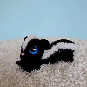 Peut inclure: Une peluche en forme de mouffette réalisée en fil noir et blanc. Ce jouet en peluche présente une bande blanche proéminente allant de la tête à la queue, de grands yeux bleus ronds avec des pupilles noires et un petit nez rose, le tout texturé.
