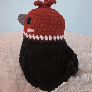 May include: A handmade crochet bird with a red head and a black body. The bird has a gray beak and a white and black patterned collar. The bird is sitting on a white, fluffy surface.