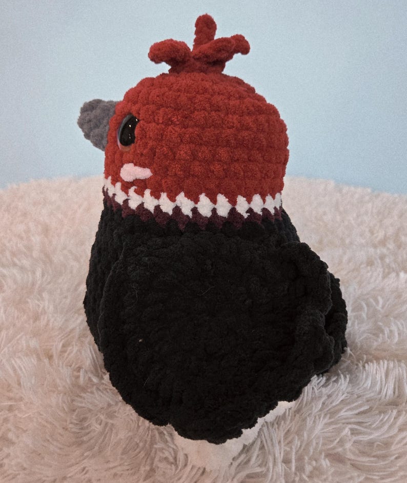 May include: A handmade crochet bird with a red head and a black body. The bird has a gray beak and a white and black patterned collar. The bird is sitting on a white, fluffy surface.