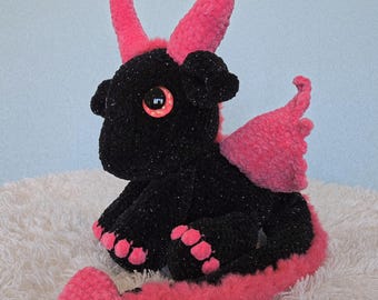 May include: A plush dragon toy crafted from black yarn with subtle glitter and bright pink yarn accents. The creature features two tall pink horns, large glossy orange eyes, pink wings, and a long pink tail with a heart-shaped tip.