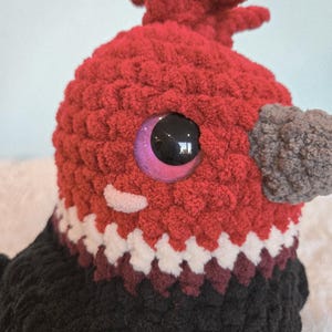 May include: Close-up of a handmade, plush bird with a red head and black body. The bird has a gray beak, large, shiny, purple-rimmed eyes, and a white and burgundy patterned collar. The bird's head has a textured, fuzzy appearance.