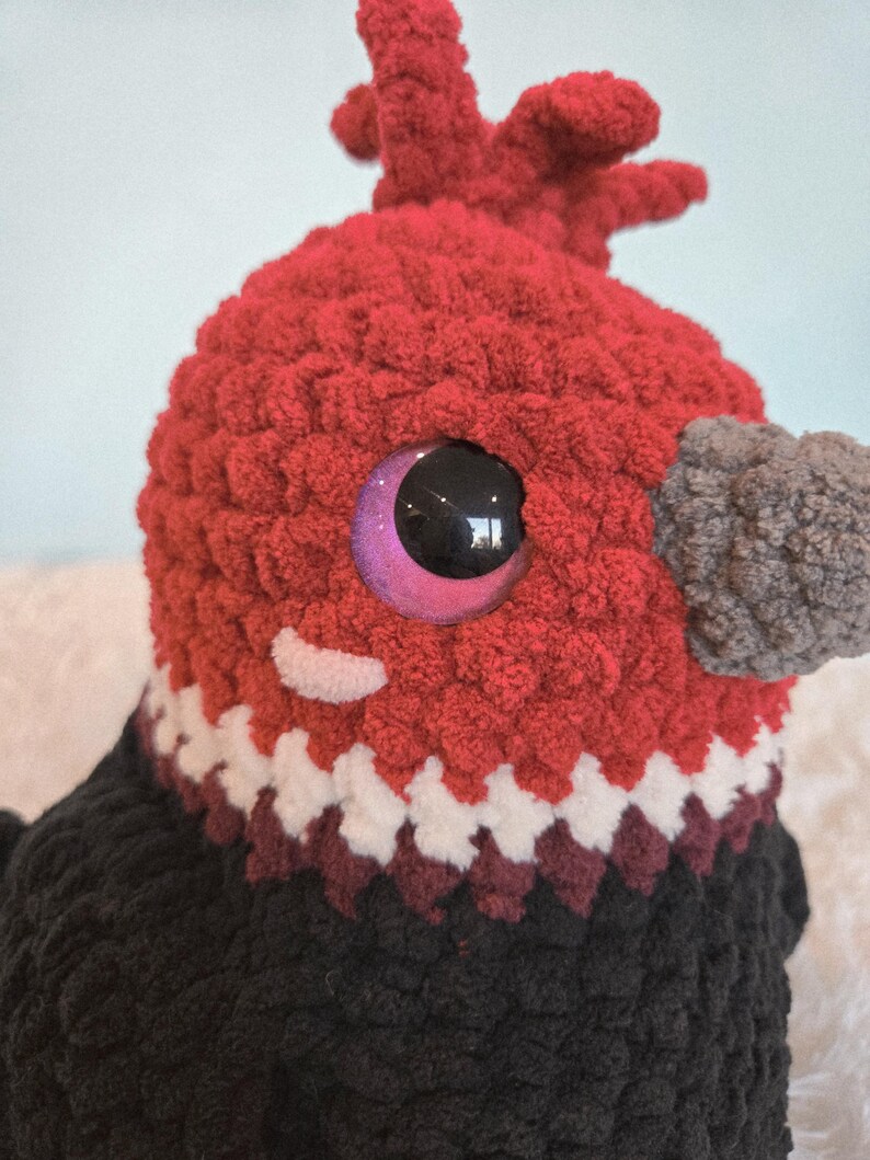 May include: Close-up of a handmade, plush bird with a red head and black body. The bird has a gray beak, large, shiny, purple-rimmed eyes, and a white and burgundy patterned collar. The bird's head has a textured, fuzzy appearance.