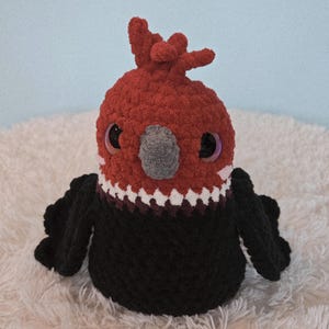 May include: A handmade, plush bird toy with a red head and crest, black body, and large, iridescent eyes. The bird has a gray beak and white and burgundy detailing around the neck. The toy is sitting on a white, fluffy surface.