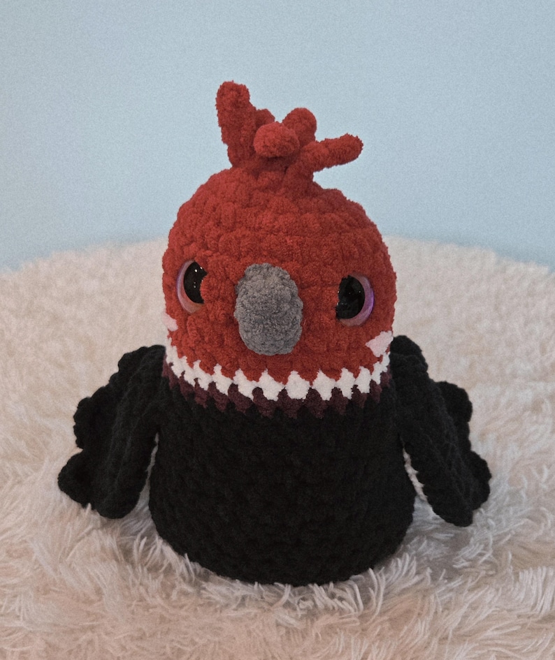 May include: A handmade, plush bird toy with a red head and crest, black body, and large, iridescent eyes. The bird has a gray beak and white and burgundy detailing around the neck. The toy is sitting on a white, fluffy surface.