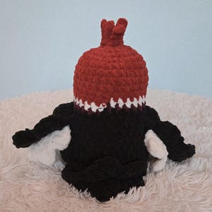 May include: A handmade crochet plush toy in the shape of a bird. The bird has a red head with a crown, a black body, and white wings. The toy is sitting on a white, fluffy surface against a light blue background.