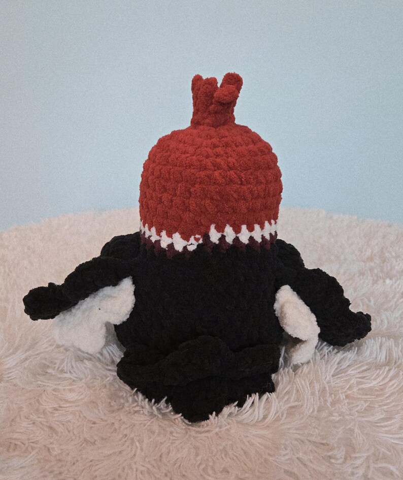 May include: A handmade crochet plush toy in the shape of a bird. The bird has a red head with a crown, a black body, and white wings. The toy is sitting on a white, fluffy surface against a light blue background.