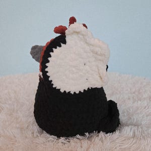 May include: A handmade crochet panda bear plushie. The panda has a black body, a white head, and red and gray accents. The plushie is sitting on a fluffy white surface against a light blue background.