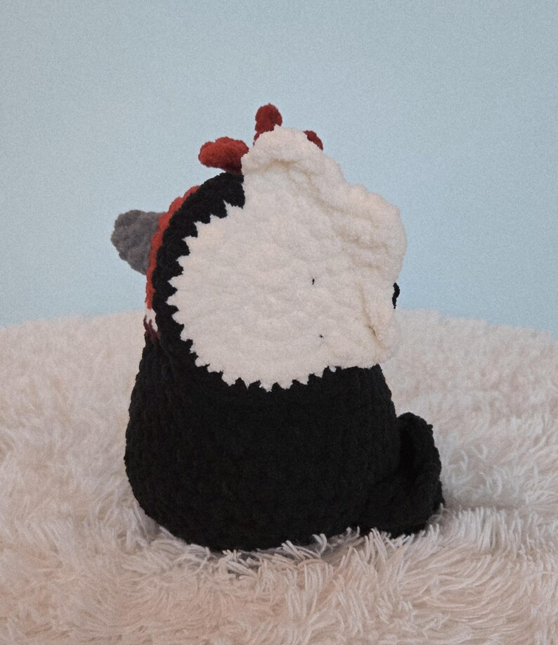 May include: A handmade crochet panda bear plushie. The panda has a black body, a white head, and red and gray accents. The plushie is sitting on a fluffy white surface against a light blue background.
