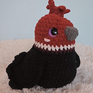 May include: A handmade crochet bird plushie with a red head, black body, and a gray beak. The bird has large, purple-rimmed eyes and a white and burgundy patterned collar. The plushie is sitting on a fluffy white surface.