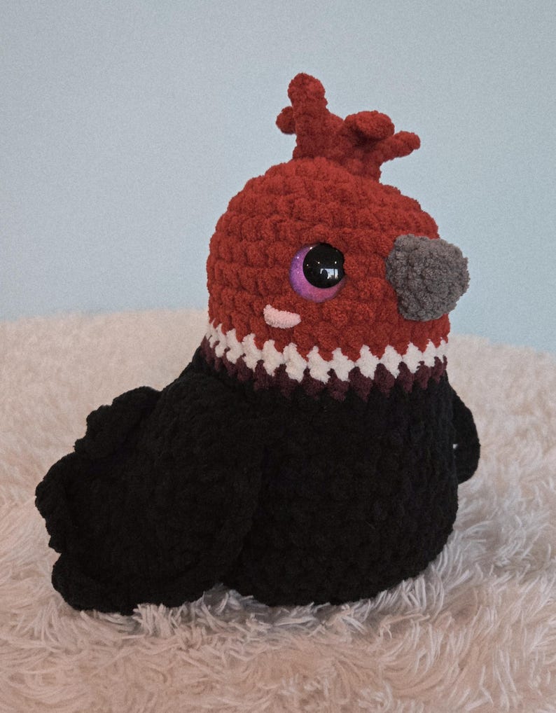 May include: A handmade crochet bird plushie with a red head, black body, and a gray beak. The bird has large, purple-rimmed eyes and a white and burgundy patterned collar. The plushie is sitting on a fluffy white surface.