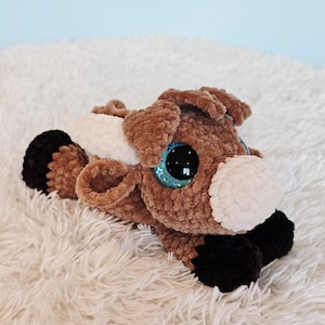 Puede incluir: Un juguete de peluche marrón y blanco con grandes ojos azules brillantes. El perro tiene patas y orejas negras. El juguete está sobre una superficie blanca y esponjosa.