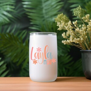 Op de afbeelding: Een witte, gematteerde glazen beker met de naam "Layla" in kleurrijke letters met bloemen en een hart.