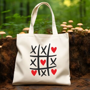 Puede incluir: Una bolsa de lona blanca con un dise&ntilde;o gr&aacute;fico en negro y rojo. El dise&ntilde;o presenta un tablero de tres en raya con corazones en lugar de las X y las O.
