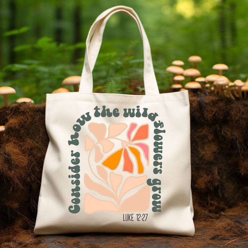 May include: A white canvas tote bag with a green, orange, and pink floral design. The text "Consider how the wild flowers grow" is written in a retro font around the flowers. The text "LUKE 12:27" is printed below the flowers.