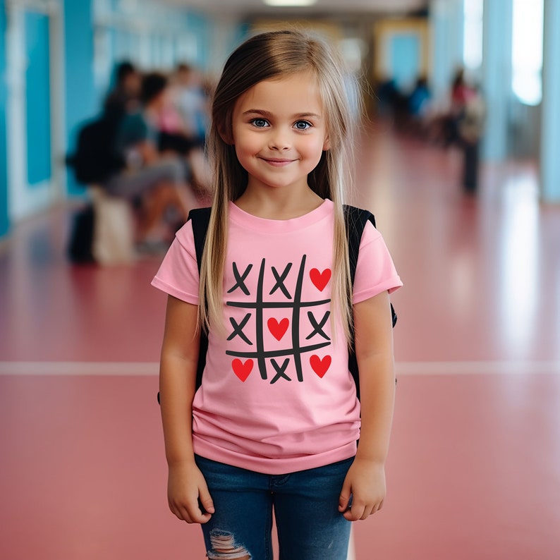 Puede incluir: Una camiseta rosa con un dise&ntilde;o de tres en raya negro y rojo. El dise&ntilde;o presenta corazones rojos y X negras.