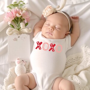 Puede incluir: Un body blanco para beb&eacute; con letras XOXO rojas y rosas con peque&ntilde;os corazones blancos. El body est&aacute; sobre una manta blanca con una diadema de flores rosas en la cabeza del beb&eacute;.