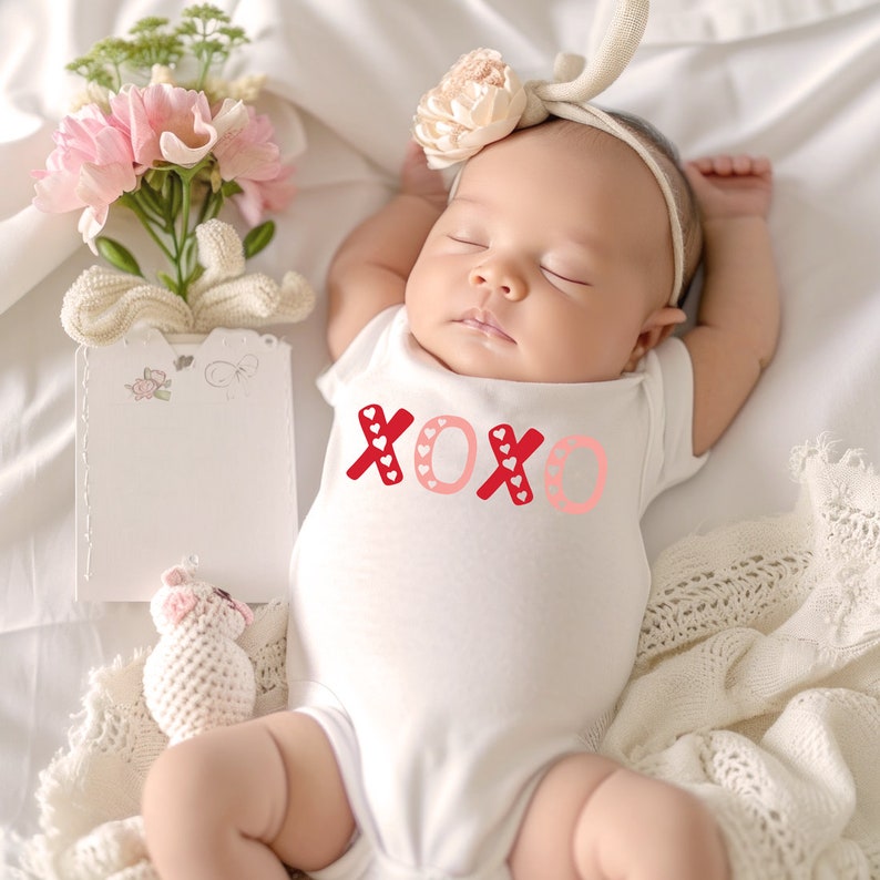 Puede incluir: Un body blanco para beb&eacute; con letras XOXO rojas y rosas con peque&ntilde;os corazones blancos. El body est&aacute; sobre una manta blanca con una diadema de flores rosas en la cabeza del beb&eacute;.