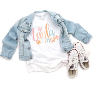 Op de afbeelding: Wit T-shirt met de naam "Layla mae" geschreven in kleurrijke letters met bloemen aan beide kanten van de naam.