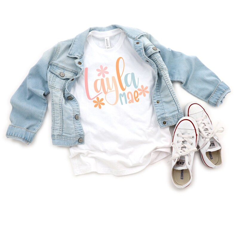 Op de afbeelding: Wit T-shirt met de naam "Layla mae" geschreven in kleurrijke letters met bloemen aan beide kanten van de naam.
