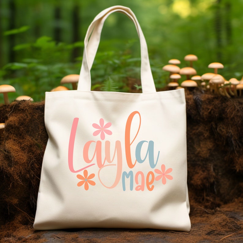Op de afbeelding: Een witte canvas tote bag met de tekst "Layla mae" in roze, oranje en blauwe letters met een bloemendesign aan beide kanten van de tekst.