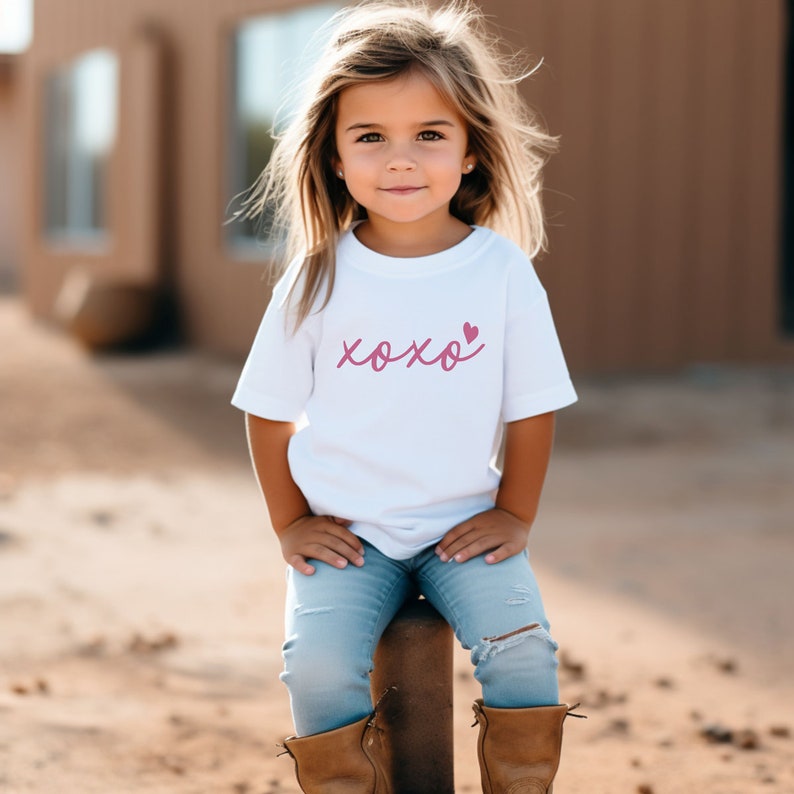 Puede incluir: Una ni&ntilde;a peque&ntilde;a con una camiseta blanca con la inscripci&oacute;n "xoxo" en rosa y un peque&ntilde;o coraz&oacute;n rosa. Lleva pantalones vaqueros azules y botas marrones.