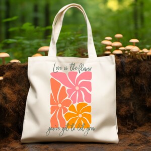May include: A white canvas tote bag with the text "Love is the flower you've got to let grow" and a graphic of three stylized flowers in pink, orange, and yellow.