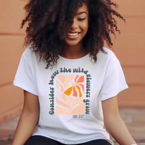 May include: White t-shirt with a graphic of colorful flowers and the text "Consider how the wild flowers grow" with the scripture reference "LUKE 12:27".