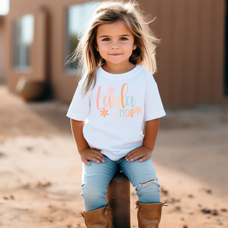 Op de afbeelding: Een wit T-shirt met de tekst "Layla mae" in kleurrijke letters met bloem accenten. Het shirt wordt gedragen door een jong meisje met lang bruin haar. Ze zit op een bruine paal en draagt ​​blauwe jeans en bruine laarzen.