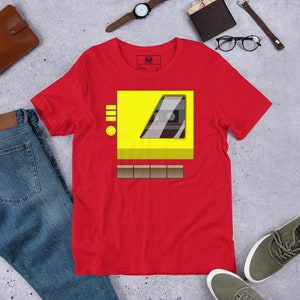 May include: Red t-shirt with a yellow cassette tape graphic. The cassette tape has a brown button and a clear window showing a black cassette tape inside.