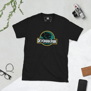 Puede incluir: Camiseta negra con un gráfico verde y amarillo de un pez prehistórico con el texto "Devonian Park" en él.