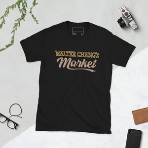 Puede incluir: Camiseta negra con un gráfico dorado que dice "Walter Chang's Market" en una fuente de estilo vintage.