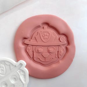 May include: A pink, circular mould featuring a dog's face wearing a firefighter's helmet. The helmet has a paw print emblem. A white plastic cutter with the same dog face design is next to the mould. The design is for a children's theme.